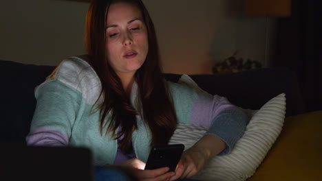 Mujer-Sentada-En-Un-Sofá-En-Casa-Por-La-Noche-Transmitiendo-O-Viendo-Una-Película-O-Programa-En-Una-Computadora-Portátil-Y-Navegando-Por-Internet-En-El-Teléfono-2